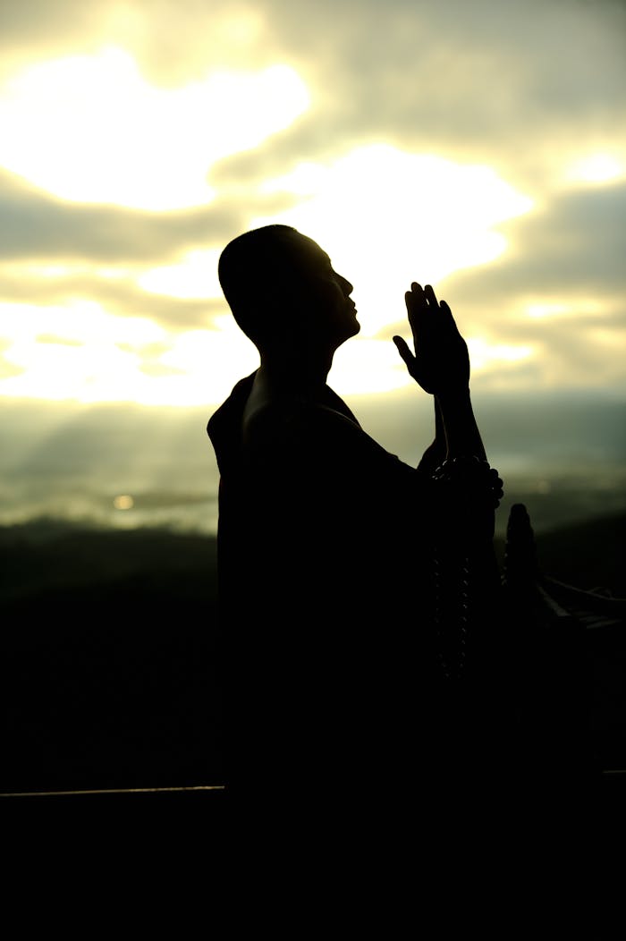 Silhouette of a person praying with hands together during sunset. Captured outdoors with a dramatic sky.