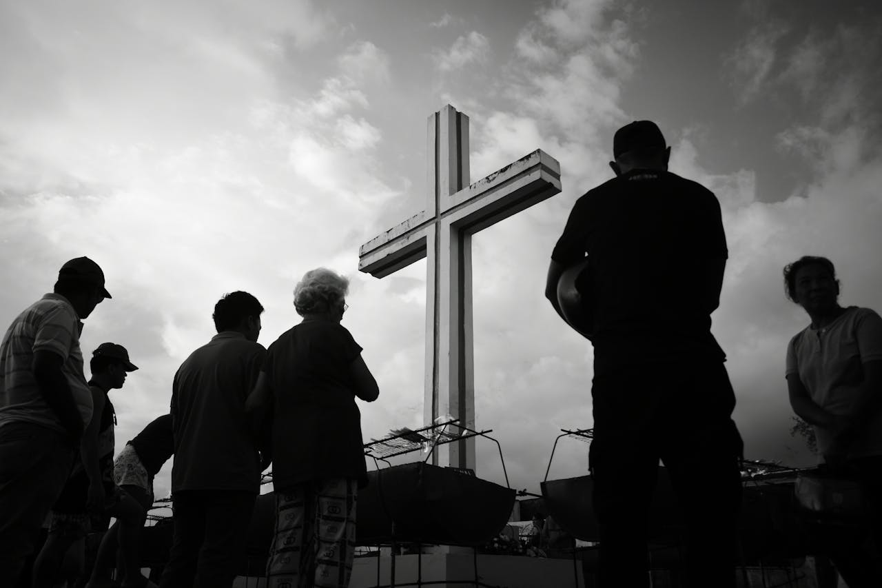 People gathered in silhouette at a large outdoor cross under dramatic skies.